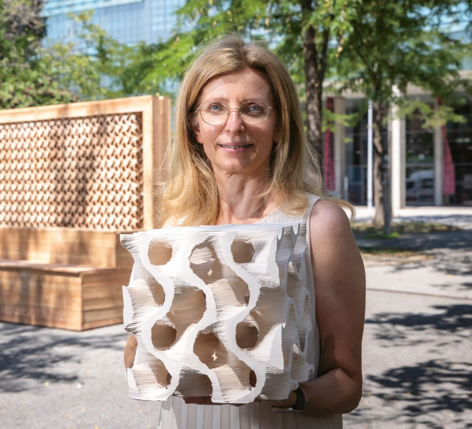 Eine Frau mit Brille hält ein weißes, skulpturales Objekt mit geometrischen Löchern in der Hand und steht im Freien in der Nähe von Bäumen und modernen Holzkonstruktionen.
