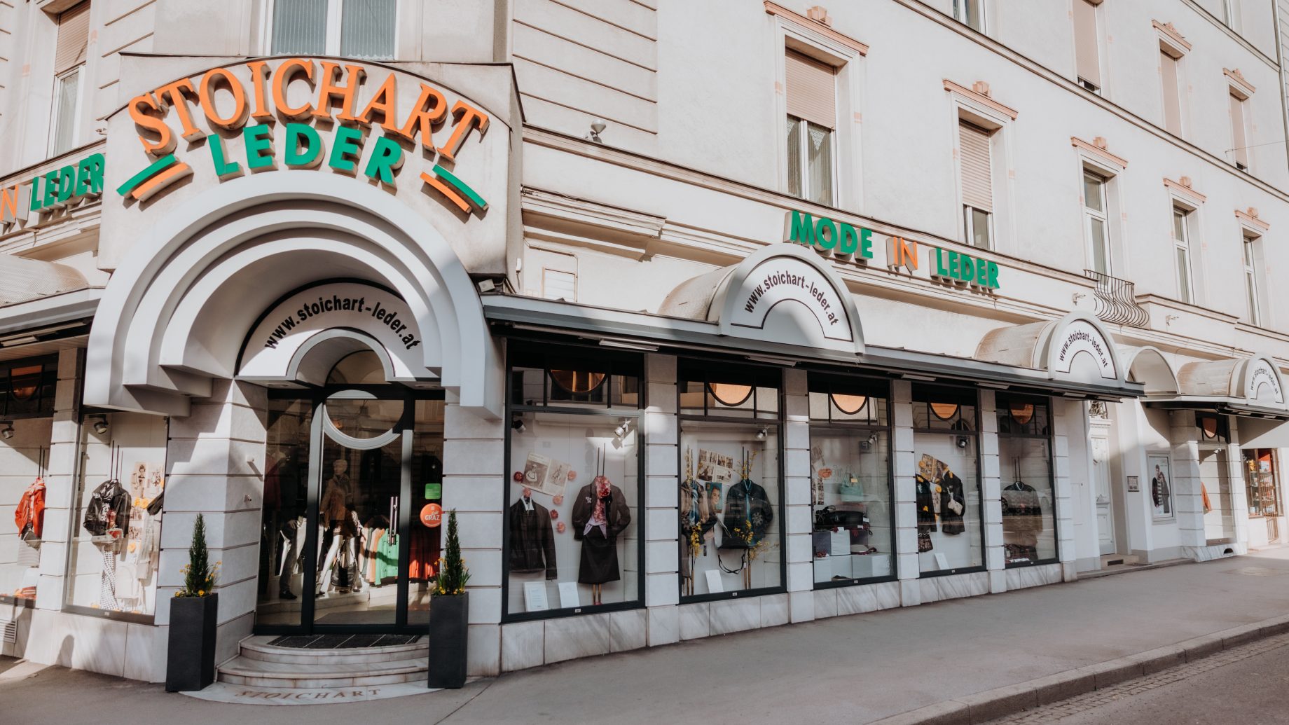 Straßenansicht eines weißen Gebäudes mit den Schildern „STOICHART LEDER“ und „MODE IN LEDER“ über den Schaufenstern, in denen Lederwaren ausgestellt sind.