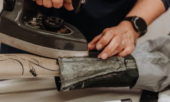 Eine Person bügelt Stoff mit einem alten Bügeleisen und drückt dabei auf einen gemusterten Stoff; ihre Hände sind zu sehen, an einer davon trägt sie eine schwarze Armbanduhr.