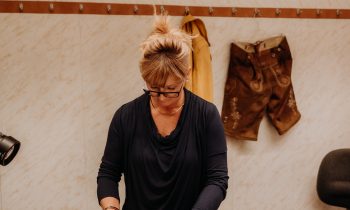 Eine Frau mit Brille arbeitet an einem Tisch in einer Nähstube an einem Stoff, im Hintergrund sind Werkzeuge und aufgehängte Kleidungsstücke zu sehen.