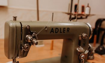 Eine grüne Adler-Nähmaschine auf einem Holztisch, mit Stoff unter der Nadel und Garnspulen im Hintergrund.