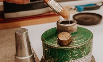 Eine Hand taucht einen Pinsel in einen kleinen grünen Metallbehälter, daneben steht ein Metallbecher auf einer Arbeitsfläche.