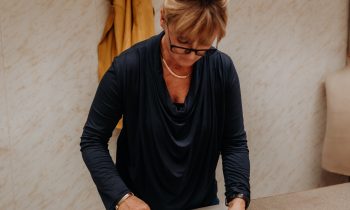 Eine Frau mit blonden Haaren, die zu einem Dutt zusammengebunden sind, arbeitet in braunen Lederhosen an einem Tisch in einer Werkstatt.