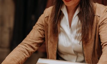 Eine Frau in einer braunen Jacke lächelt, während sie eine Schachtel öffnet, in der sich ein gelbes Kleidungsstück und eine Karte mit der Aufschrift „Willkommen bei …“ befinden.