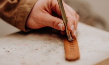 Eine Person prägt mit einem Metallwerkzeug und einem Hammer auf einer Steinoberfläche Buchstaben in ein Lederband ein.