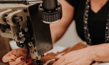 Eine Person mit roten Fingernägeln näht mit einer Nähmaschine braunen und weißen Stoff auf einem weißen Tisch fest.