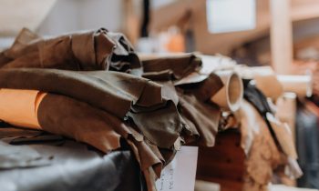 Auf einem Holzregal in einer Werkstatt liegen gestapelte Lederrollen in verschiedenen Farben, wobei an einigen der Materialien handgeschriebene Etiketten angebracht sind.