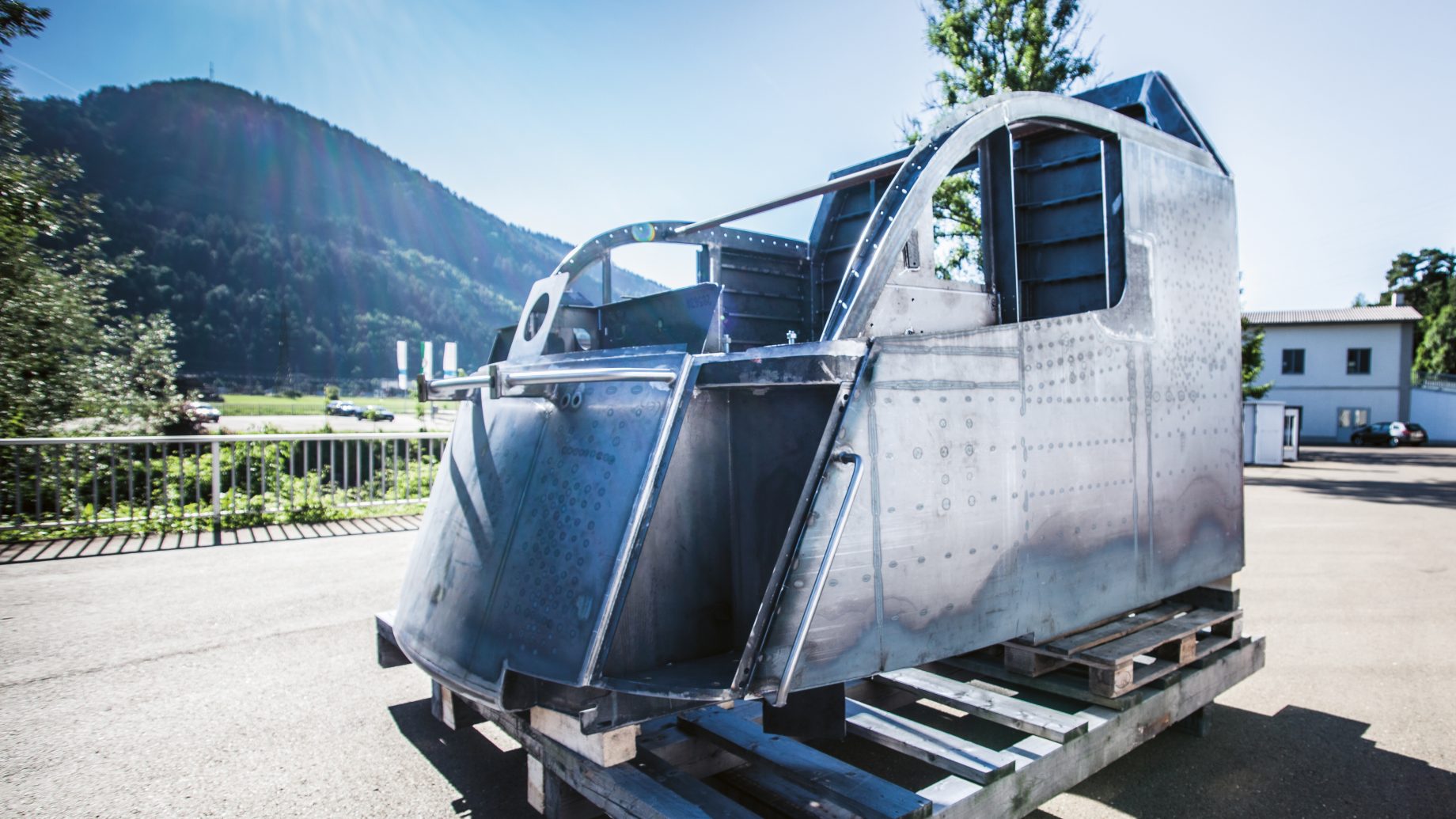 Ein metallener Cockpitrahmen steht im Freien auf einer Holzpalette; im Hintergrund sind unter einem klaren Himmel Berge, Bäume und Gebäude zu sehen.