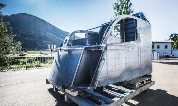 Ein metallener Cockpitrahmen steht im Freien auf einer Holzpalette; im Hintergrund sind unter einem klaren Himmel Berge, Bäume und Gebäude zu sehen.