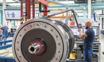 Ein Mann bedient in einer Fabrik eine Maschine neben einem großen Zugrad; um ihn herum sind Computerbildschirme und Geräte zu sehen.