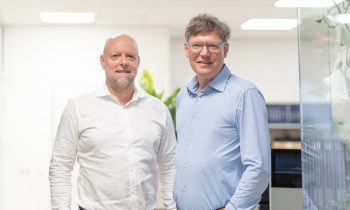 Zwei Männer stehen in einem Büro und lächeln beide. Der eine trägt ein weißes Hemd, der andere ein hellblaues Hemd. Der Hintergrund ist hell und modern.