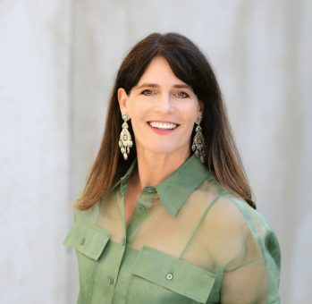 Eine Frau mit langen braunen Haaren, die eine durchsichtige grüne Bluse und große Ohrringe trägt und vor einem hellen, unscharfen Hintergrund lächelt.