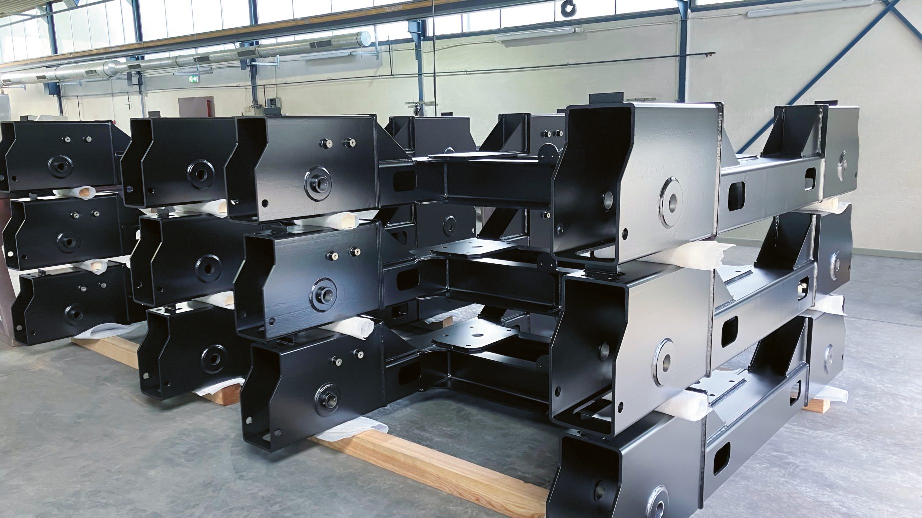 In einer Industriehalle mit Betonboden und heller Deckenbeleuchtung sind mehrere große, schwarze Metallrahmen in Reihen gestapelt.