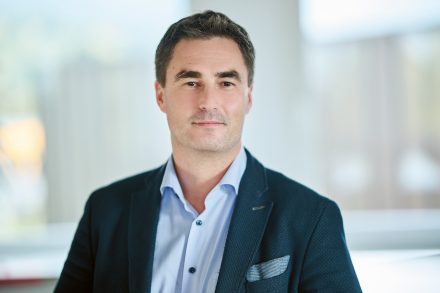Ein Mann in einem marineblauen Blazer und einem hellblauen Hemd steht in einem Raum und blickt mit ausdruckslosem Gesicht in die Kamera.