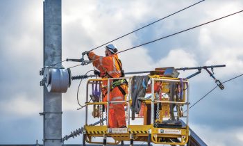 Ein Arbeiter in orangefarbener Schutzkleidung repariert unter bewölktem Himmel von einer gelben Hubarbeitsbühne neben einem Strommast aus die Freileitungen.
