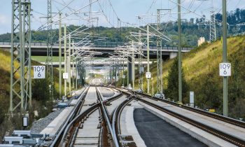 Geschwungene Bahngleise mit Oberleitungen verlaufen unter einer Brücke hindurch durch grasbewachsene Hügel, an deren beiden Seiten Schilder mit der Aufschrift „109 6“ stehen.