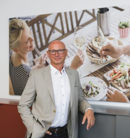 Ein Mann in einem hellen Anzug steht vor einem an der Wand hängenden Foto, auf dem drei Personen zu sehen sind, die gemeinsam essen und etwas trinken.