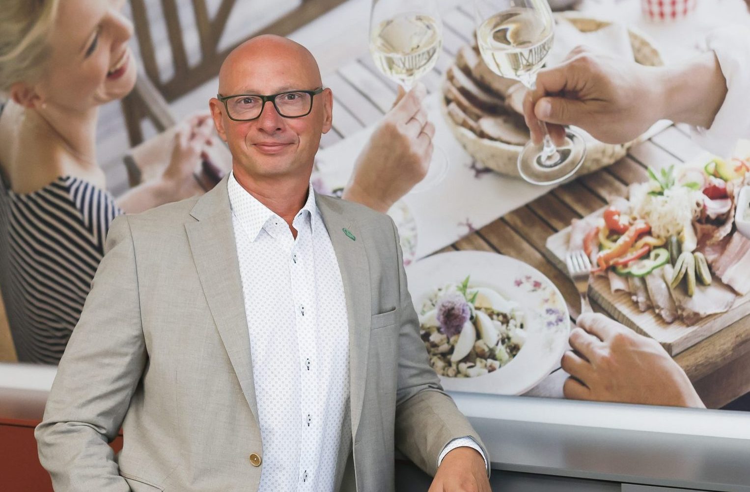Ein Mann in einem hellen Anzug steht vor einem an der Wand hängenden Foto, auf dem drei Personen zu sehen sind, die gemeinsam essen und etwas trinken.