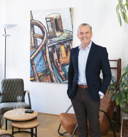 Ein lächelnder Mann in einem marineblauen Blazer steht in einem hellen, mit Pflanzen geschmückten Raum, an dessen Wänden abstrakte Kunstwerke hängen und in dem moderne Stühle um einen kleinen Tisch herum stehen.