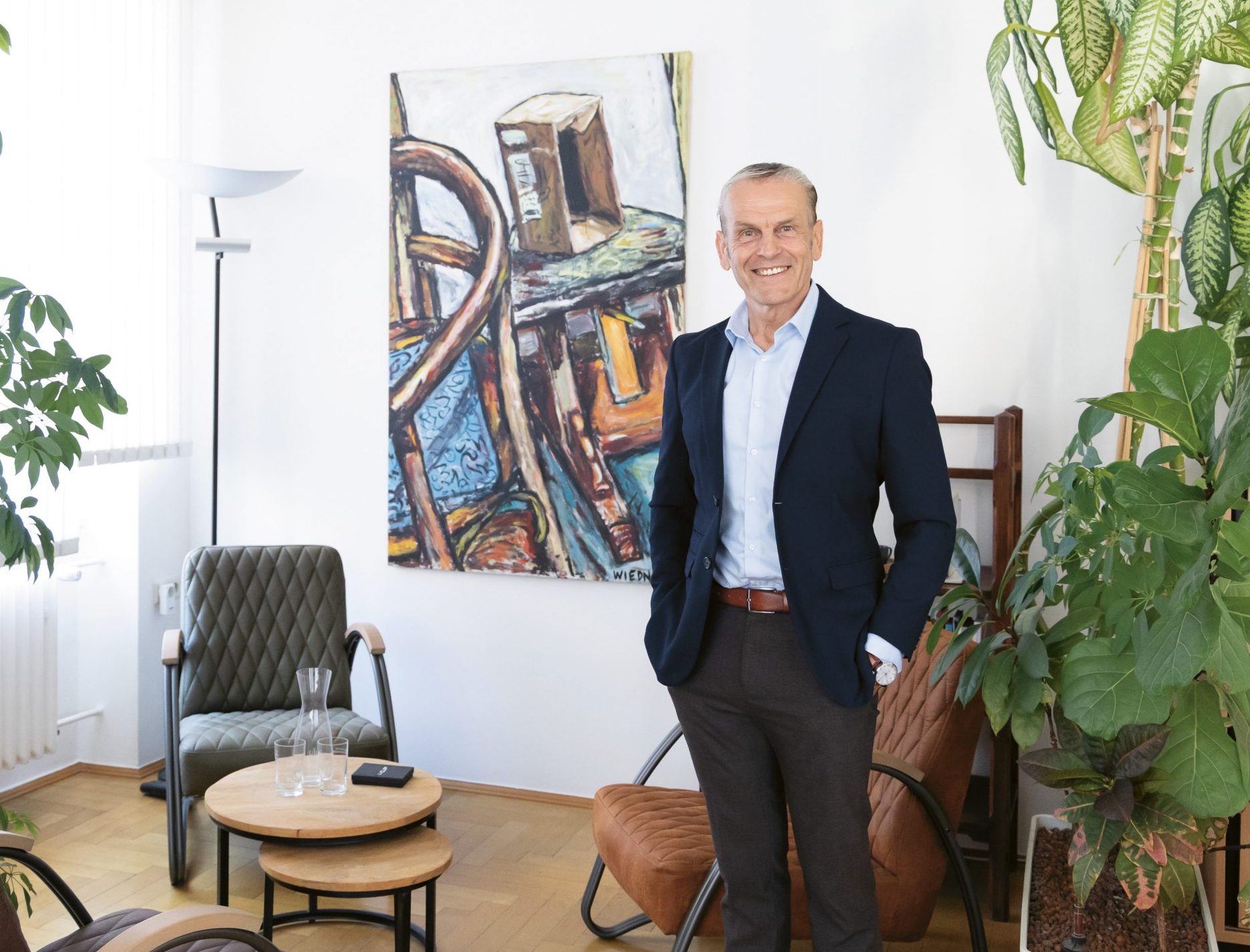 Ein lächelnder Mann in einem marineblauen Blazer steht in einem hellen, mit Pflanzen geschmückten Raum, an dessen Wänden abstrakte Kunstwerke hängen und in dem moderne Stühle um einen kleinen Tisch herum stehen.