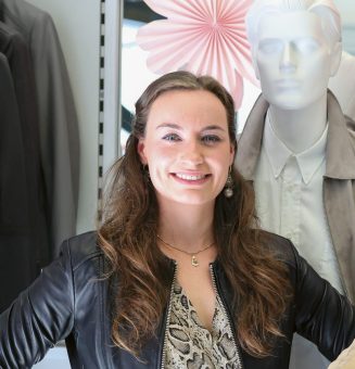 Eine Frau lächelt in einem Bekleidungsgeschäft, während sie zwischen Schaufensterpuppen steht, die Jacken und Kleider tragen, über denen rosa und weiße Zierblumen hängen.
