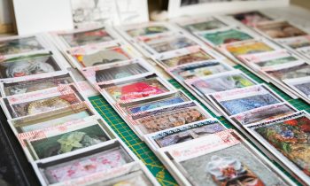 Auf einer grünen Schneidematte sind Stapel bunter Postkarten mit verschiedenen Kunstwerken ordentlich angeordnet, im Hintergrund sind einige Schwarz-Weiß-Drucke zu sehen.