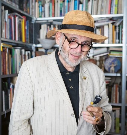 Ein älterer Mann in einem gestreiften Anzug und einem Strohhut lächelt, hält eine Brille in der Hand und steht vor einem Bücherregal, das mit Büchern gefüllt ist.