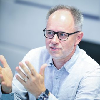 Ein Mann mit Brille und einem hellen Hemd gestikuliert mit den Händen, während er in einem Innenraum spricht.