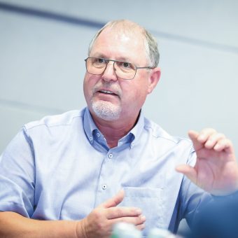 Ein älterer Mann mit Brille und kurzem grauen Bart spricht und gestikuliert dabei mit einer Hand; er sitzt in einem Raum vor einem hellen Hintergrund.