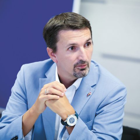 Ein Mann in einem hellblauen Anzug sitzt an einem Tisch, hat die Hände gefaltet und blickt mit nachdenklichem Gesichtsausdruck zur Seite.