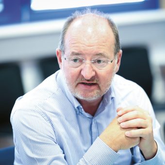 Ein Mann mit Brille und Bart, der ein hellblaues Hemd trägt, sitzt in einem Raum, die Hände gefaltet, und scheint sich zu unterhalten.