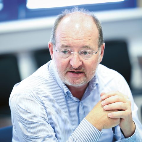 Ein Mann mit Brille und Bart, der ein hellblaues Hemd trägt, sitzt in einem Raum, die Hände gefaltet, und scheint sich zu unterhalten.