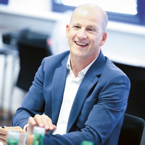 Ein lächelnder Mann in einem blauen Anzug sitzt an einem Tisch, vor dem Flaschen stehen, und hält einen Stift in der Hand – in einer hellen, modernen Büroumgebung.