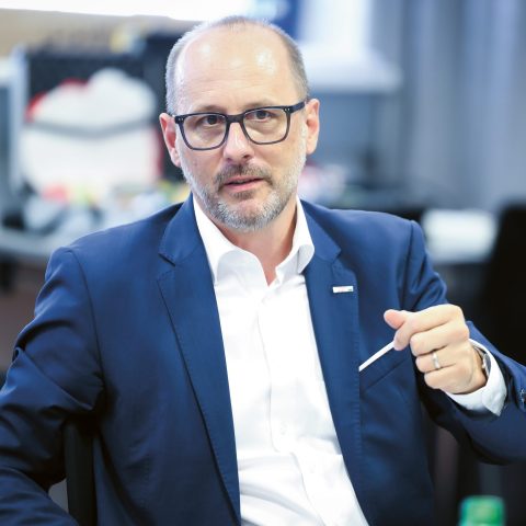Ein Mann in Anzug und Brille sitzt in einem modernen Büro an einem Schreibtisch und gestikuliert mit einem Stift.