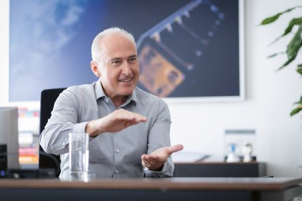 Ein lächelnder Mann in einem grauen Hemd gestikuliert mit den Händen, während er an einem Schreibtisch sitzt, auf dem ein Glas Wasser, Bücher und eine Pflanze stehen.