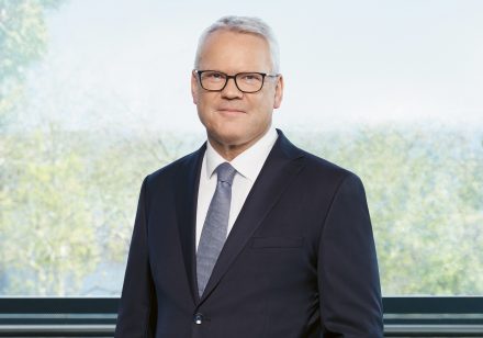Ein Mann in Anzug und Krawatte steht vor einem Fenster, durch das man Bäume und den Himmel sehen kann. Er hat kurzes graues Haar und trägt eine Brille.