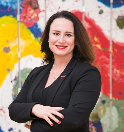 Eine Frau in einem schwarzen Blazer mit verschränkten Armen, die vor einem farbenfrohen Hintergrund mit abstrakten Kunstmotiven lächelt.