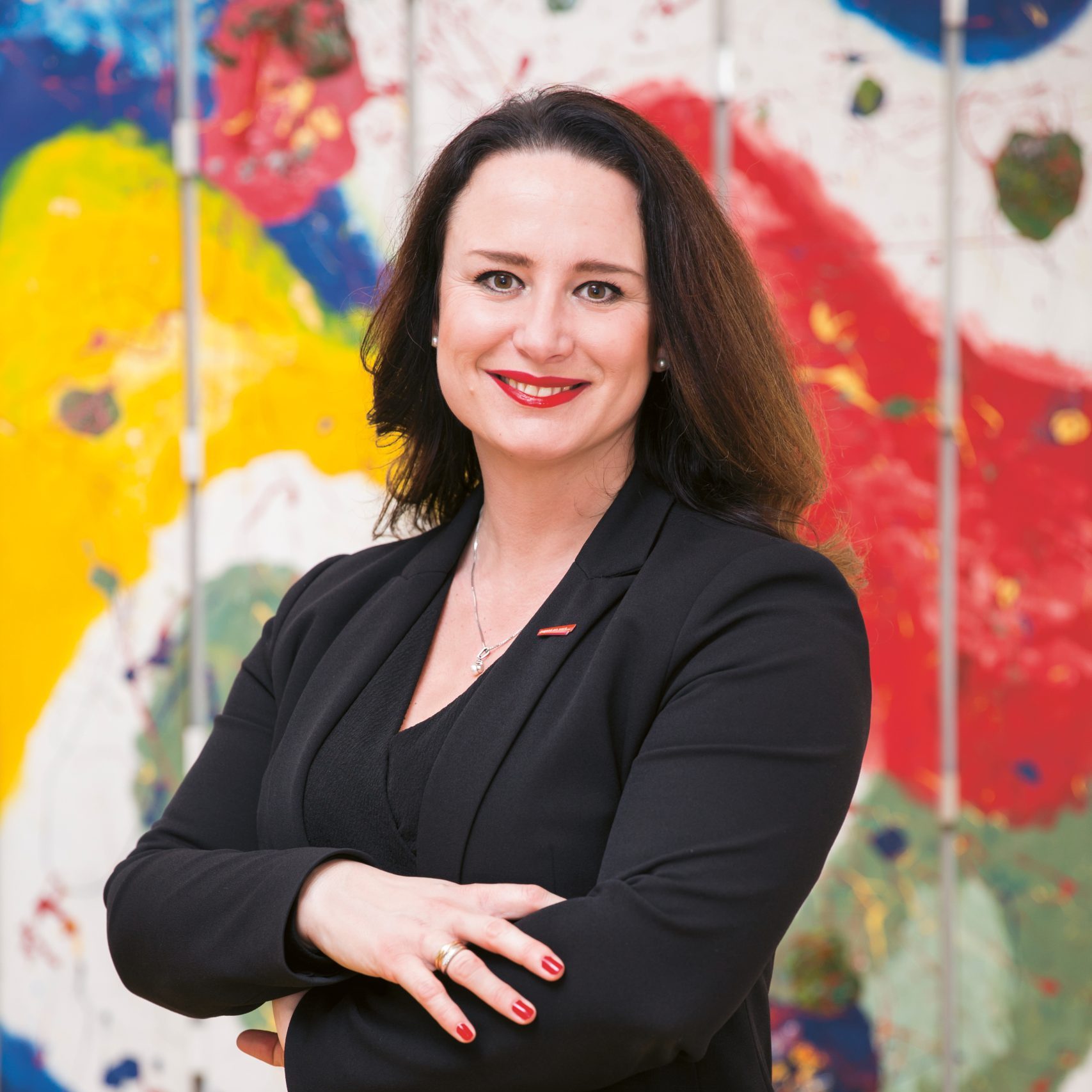 Eine Frau in einem schwarzen Blazer mit verschränkten Armen, die vor einem farbenfrohen Hintergrund mit abstrakten Kunstmotiven lächelt.