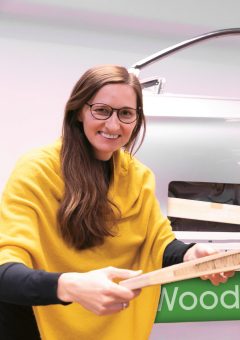 Eine Frau mit langen braunen Haaren und Brille lächelt, während sie ein Holzbrett in der Hand hält; sie trägt ein gelbes Oberteil und steht neben einem grün-weißen Schild.