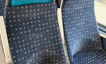 Zwei leere Zugsitze mit blauer und türkisfarbener Polsterung mit Punktmuster, die neben einem Fenster stehen, durch das man einen anderen Zug sehen kann.