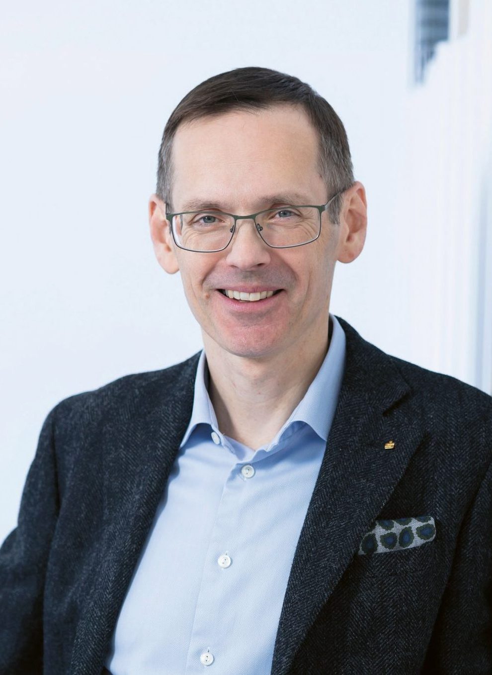 Ein Mann mit kurzen braunen Haaren und Brille lächelt; er trägt ein blaues Hemd, einen dunklen Blazer und ein gemustertes Einstecktuch und befindet sich in einem hellen Innenraum.