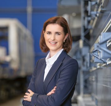 Eine Frau in einem marineblauen Kostüm steht lächelnd mit verschränkten Armen neben Industrieanlagen auf Bahngleisen in einer modernen Anlage.