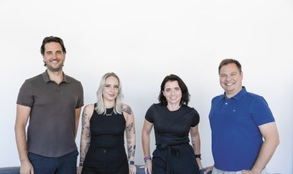 Vier Personen stehen nebeneinander, lächeln und tragen Freizeitkleidung vor einem schlichten weißen Hintergrund.