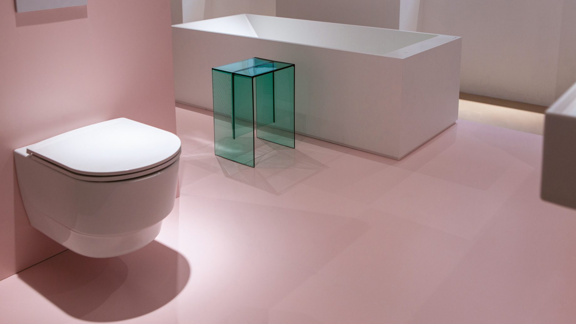 Ein an der Wand montiertes weißes WC, eine weiße rechteckige Badewanne und ein durchscheinender, blaugrüner Beistelltisch in einem Raum mit rosa Bodenbelag.
