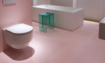 Ein an der Wand montiertes weißes WC, eine weiße rechteckige Badewanne und ein durchscheinender, blaugrüner Beistelltisch in einem Raum mit rosa Bodenbelag.