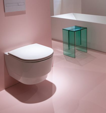 Ein an der Wand montiertes weißes WC, eine weiße rechteckige Badewanne und ein durchscheinender, blaugrüner Beistelltisch in einem Raum mit rosa Bodenbelag.
