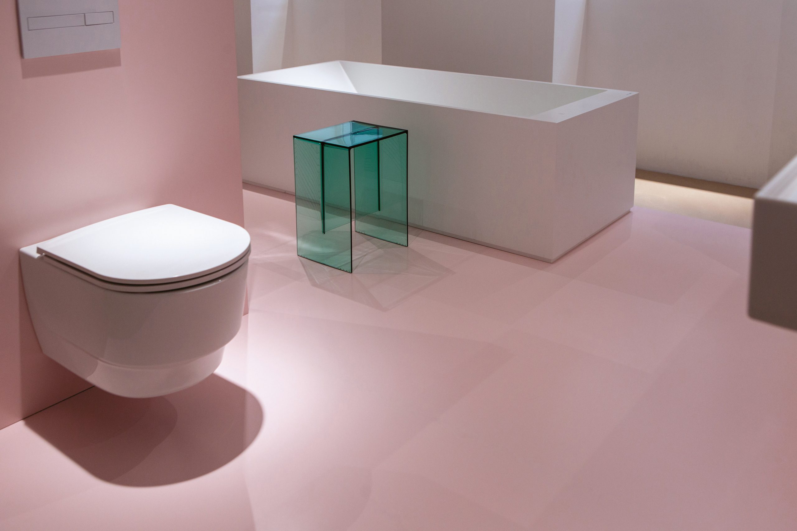 Ein an der Wand montiertes weißes WC, eine weiße rechteckige Badewanne und ein durchscheinender, blaugrüner Beistelltisch in einem Raum mit rosa Bodenbelag.
