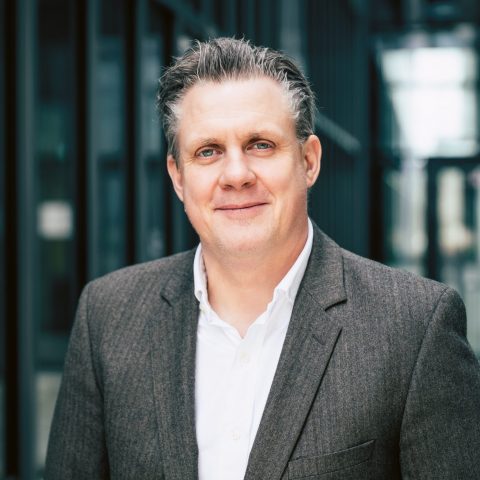 Ein Mann mit kurzem grauem Haar, der einen braunen Blazer und ein weißes Hemd trägt und im Freien in der Nähe von Glasgebäuden lächelt.