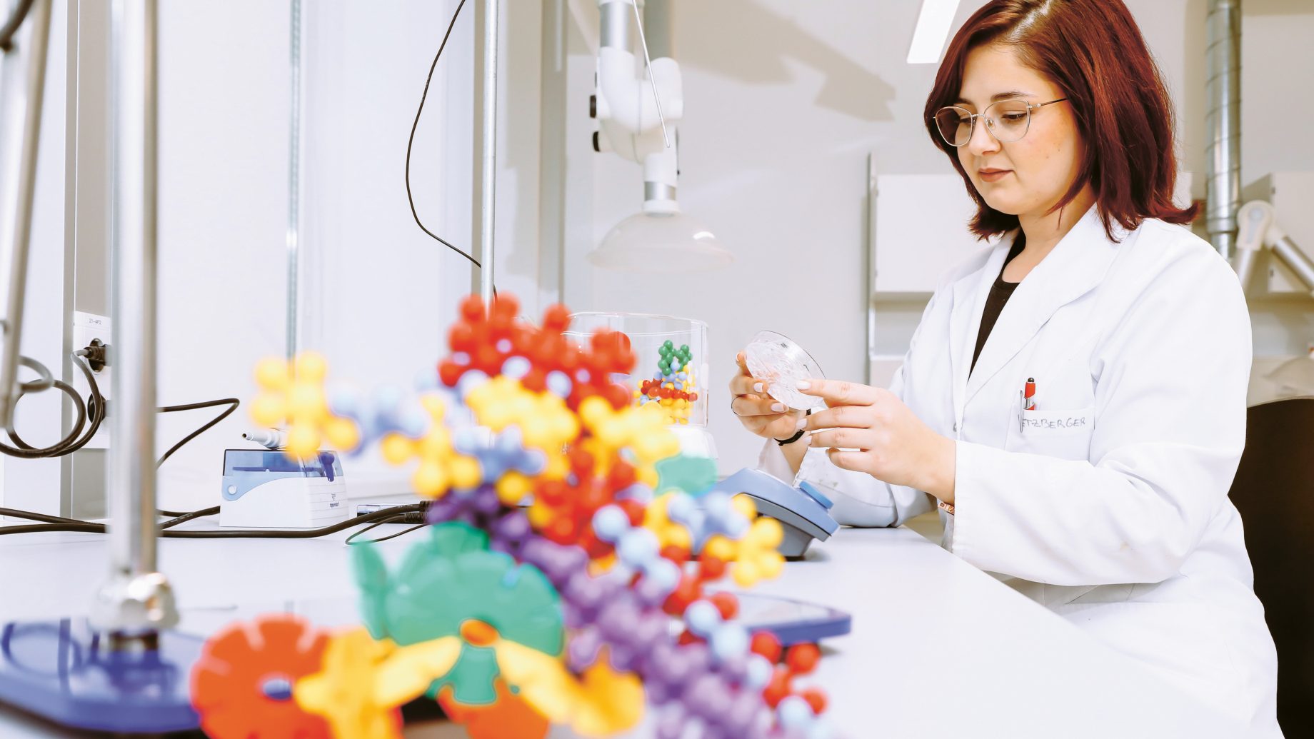 Ein Wissenschaftler im Laborkittel, der neben einem bunten Molekülmodell auf einem Labortisch eine Petrischale untersucht.
