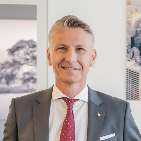 Ein lächelnder Mann in einem grauen Anzug und einer roten Krawatte mit Polka-Punkten steht neben einem Schreibtisch in einem modernen Büro, an dessen Wänden gerahmte Fotos hängen.